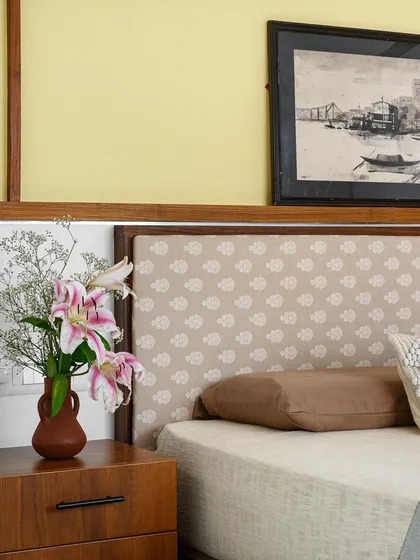 A close-up of the guest bedroom in 'Poorna', highlighting the fabric-upholstered headboard and the crisscrossing wooden ledges on the yellow accent wall.
