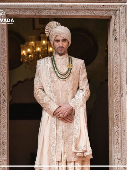 A groom in a Shauhar sherwani, framed by a majestic doorway. The rich embroidery and the emerald green necklace create a look of unparalleled royal splendor.