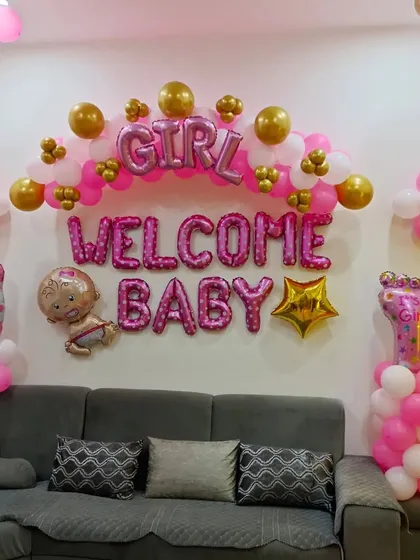 A cheerful and bright welcome home decoration for a baby girl. This setup is perfect for an apartment living room, featuring "WELCOME BABY" and "GIRL" foil balloons, a pink and gold balloon arch, and cute baby-themed foil characters.