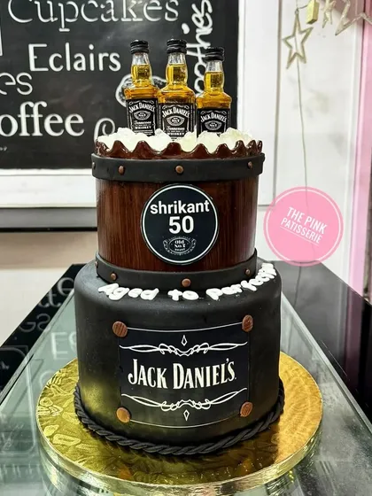 A fun and creative two-tiered cake designed for a Jack Daniel's enthusiast's 50th birthday. The top tier is crafted to look like a wooden whiskey barrel, complete with miniature liquor bottles, while the bottom tier features the iconic branding.