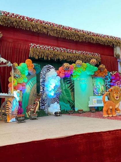 A full stage view of a farmhouse jungle party, with a red curtain backdrop, multiple animal props, and vibrant balloon decorations.