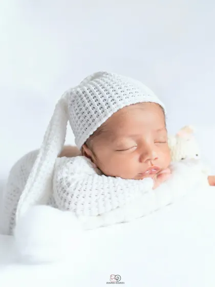 A pure and simple newborn portrait against a clean white background. The baby is dressed in a white knitted outfit and sleepy hat, showcasing their angelic features.