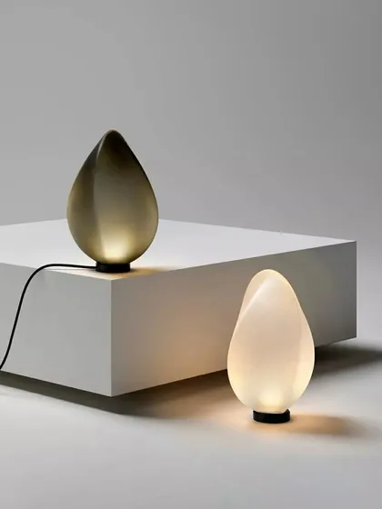 The Solace table lamp by Ross Gardam is presented in two distinct forms, one a luminous white and the other a smoky grey. These sculptural glass pieces, shaped like luminous tears, exemplify my curation of smaller, artistic objects that provide both function and beauty for intimate settings.