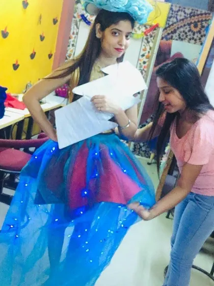 A student fits a creative, weather-themed garment on a fellow student. The design, which was developed from a sketch, features a skirt with embedded LED lights and a lightning bolt motif, showcasing innovation and teamwork.