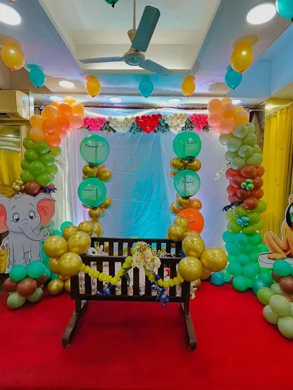 A unique animal-themed naming ceremony (Barsa). The setup features a decorated wooden cradle (palna) surrounded by friendly jungle animal cutouts like a lion and elephant, and a colorful balloon backdrop, making it a memorable day.