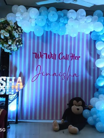 A naming ceremony decoration announcing the name "Jinaisha". The setup features a striped backdrop, a blue and white balloon arch, and light-up letters spelling out the new baby's name.