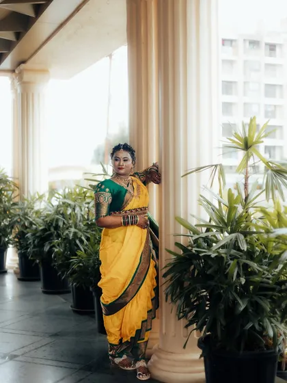 A beautiful portrait of a Maharashtrian bride in her yellow Nauvari saree, standing elegantly amongst pillars and greenery.
