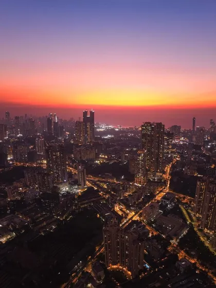 A high-altitude drone shot of Mumbai after sunset, with the city lights beginning to glow under a fiery orange sky.