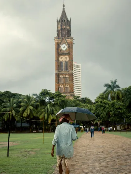 A person in traditional attire walks with an umbrella at Oval Maidan. This image is a beautiful blend of culture, history (the clock tower), and the timelessness of the monsoon.