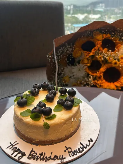 A simple and elegant blueberry cheesecake with a handwritten "Happy Birthday" message, paired with a bouquet of sunflowers in the background.