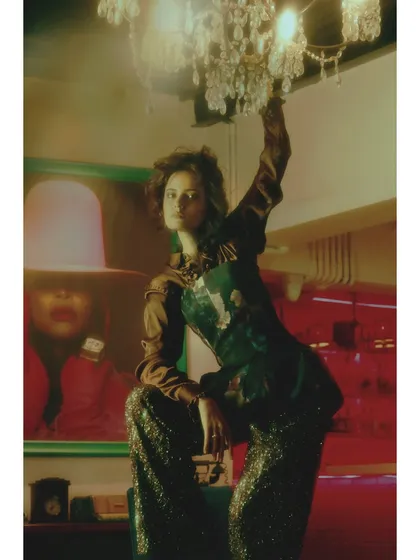 From 'Chapter 7: The Future is Daring'. The styling becomes bold and eclectic, mixing a satin blouse with sparkling sequin trousers. The pose is assertive, reaching for a chandelier, symbolizing reaching for a brighter future.