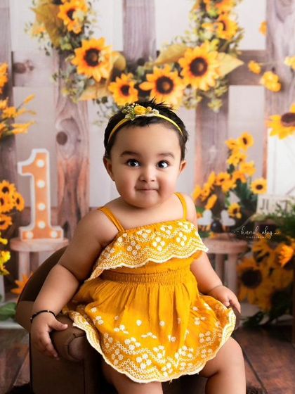 A baby girl with a sweet smile poses for her sunflower-themed first birthday portrait.