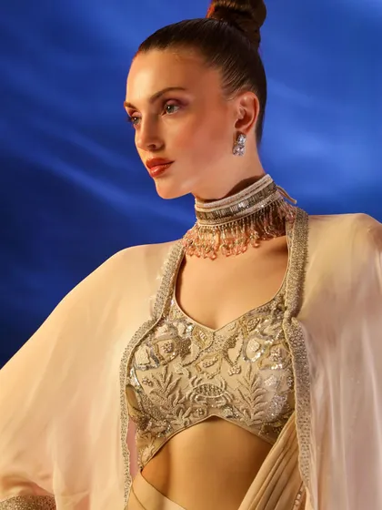 A regal shot of a model in a beige cape-and-saree ensemble. The high-neck choker and elegant updo complete this sophisticated, high-fashion ethnic look.