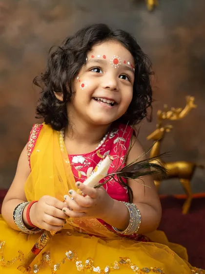 A beautiful little girl dressed as Radha for a Janmashtami themed shoot, holding a peacock feather with a joyful smile.