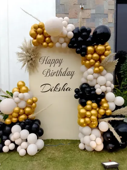 An outdoor birthday decoration with a custom backdrop and a balloon arch in black, white, and gold, accented with pampas grass.