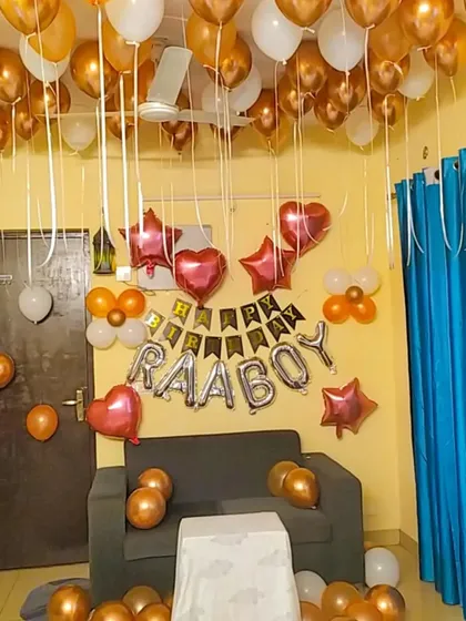 A birthday decoration for a 'Raaboy' using gold, white, and orange balloons. The setup includes star and heart-shaped foil balloons and a 'Happy Birthday' banner for a personal touch.
