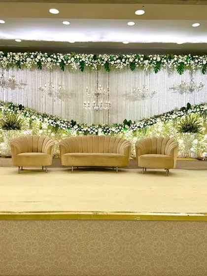 An elegant stage decor featuring a curtain of white florals and multiple crystal chandeliers. The neutral-toned seating and lush greenery create a look of understated luxury.