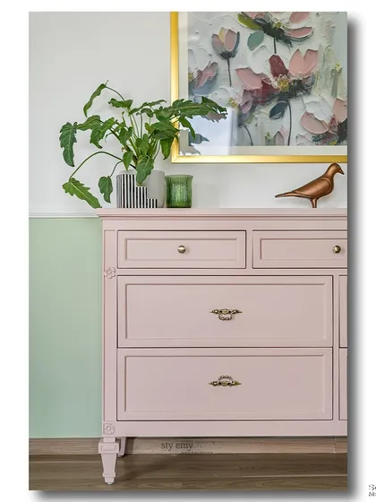 A charming pastel pink dresser set against a two-toned wall. The floral artwork above and the classic hardware give this piece a vintage feel, perfect for adding a soft, romantic touch to a room.