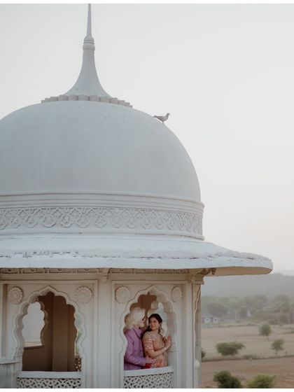 A fairytale view from a palace turret in Jaipur. The couple shares a quiet moment, framed by the window, with a lone bird perched on the dome, adding a touch of serendipity to this romantic pre-wedding scene.