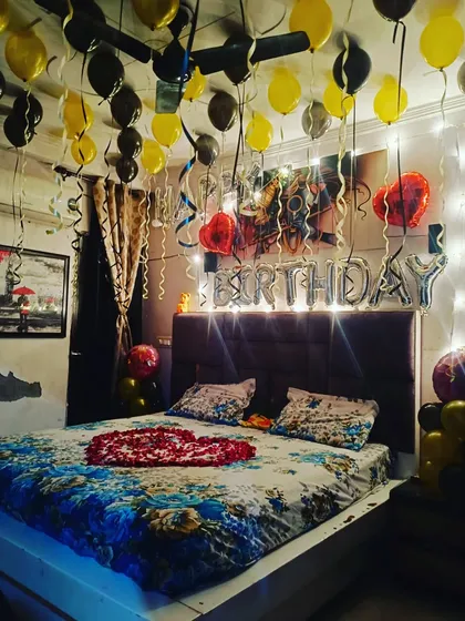 A birthday room decoration featuring yellow and black balloons and a heart made of rose petals on the bed. The fairy lights add a warm and cozy glow to the room.