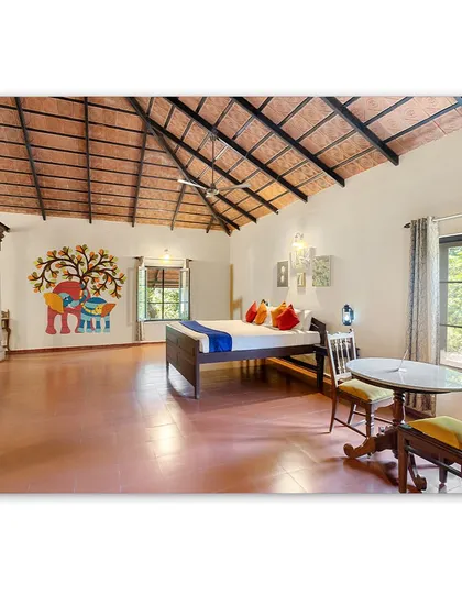 The interior of a cottage at Magnolia Resorts, featuring a spacious room with a high, exposed-beam ceiling and colorful Gond art on the wall. The design is a blend of rustic charm and local artistry.