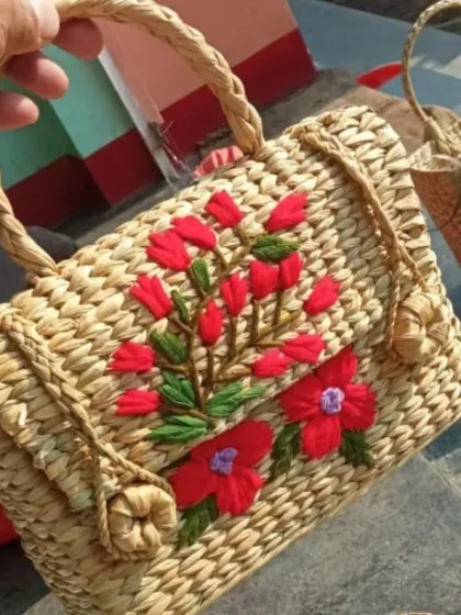A close-up view of a handwoven bag, highlighting the intricate weave of the natural fibers and the vibrant red floral embroidery. The braided handle adds to its rustic charm, showcasing the detailed work of our artisans.