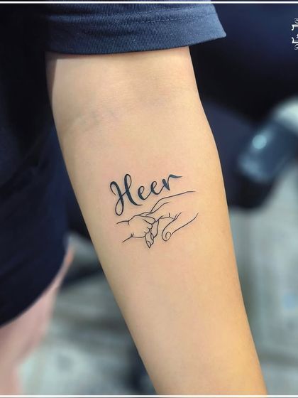 A custom tattoo combining the name "Heer" with a line art drawing of a parent and child holding hands. This is a deeply personal and emotional design that we were honored to create.