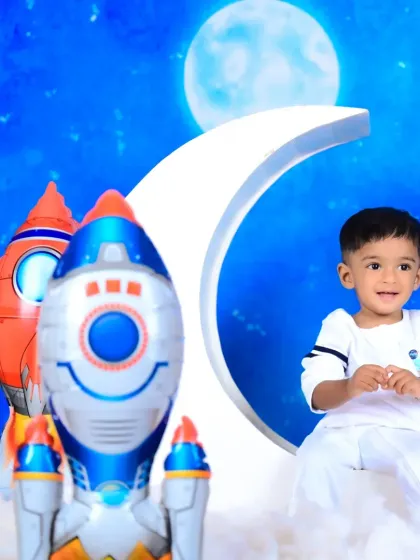 A baby boy dressed as an astronaut, sitting on a moon prop with rocket balloons.