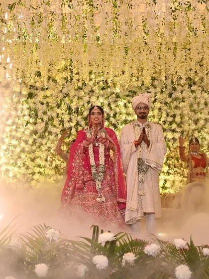 The couple stands together after the Jaimala, enveloped in a mystical cloud of smoke with a single pyrotechnic sparkler. The backdrop of white flowers adds to the ethereal beauty of the scene.