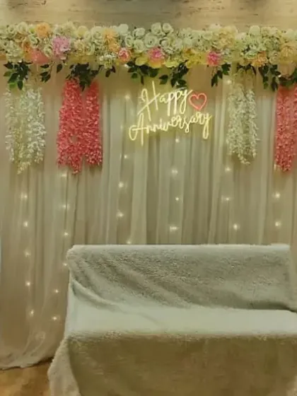 A beautiful floral backdrop for a 50th wedding anniversary. The setup includes a 'Happy Anniversary' neon sign, hanging wisteria flowers, and large '50' marquee numbers.