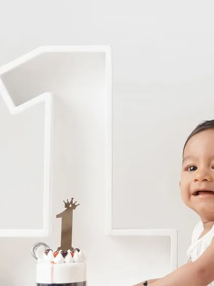 A close-up shot of a smiling baby boy next to his first birthday cake, which is topped with a '1' and a crown.
