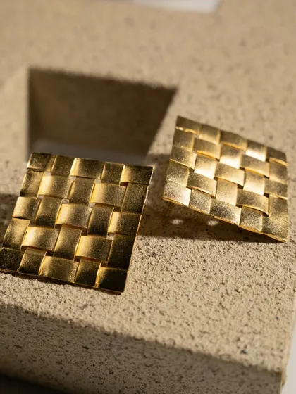 A product shot of textured gold earrings. The directional light creates beautiful highlights and shadows, emphasizing the woven pattern and making the jewelry look luxurious.