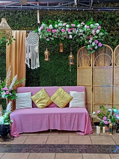 A cozy and chic boho-themed seating area for a relaxed party. The setup includes a green hedge wall, a dreamcatcher, and comfortable seating with plush cushions.
