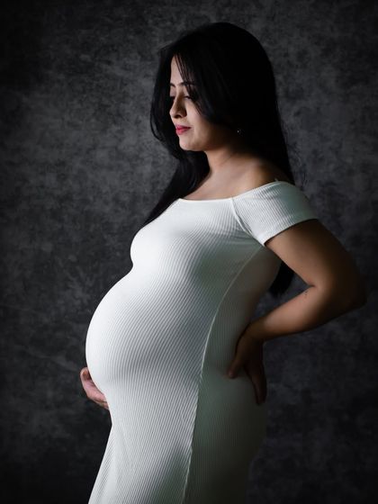 A serene and classic black and white portrait. The soft studio lighting beautifully sculpts the mom-to-be's form in a simple white dress.