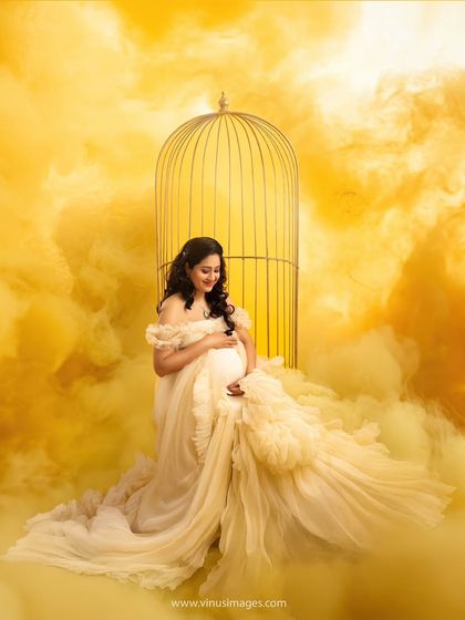 A dreamy, ethereal portrait. The mother-to-be, in a soft yellow gown, is surrounded by a golden mist and a birdcage prop, suggesting a precious life being protected and cherished.