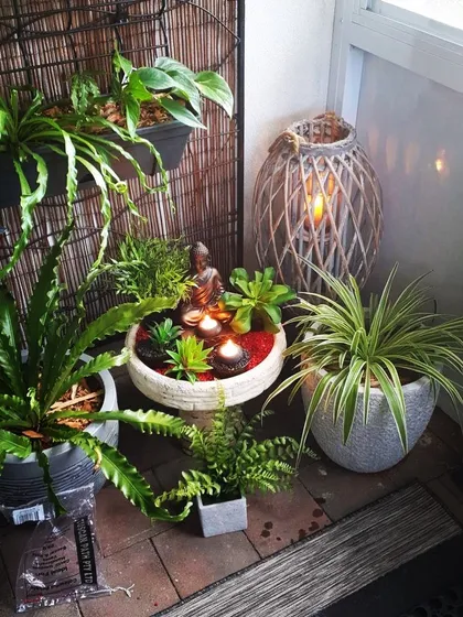 This tranquil corner features a miniature zen garden with a Buddha statue and floating candles. The arrangement is surrounded by various potted plants and a woven lantern, creating a perfect spot for meditation and peace.