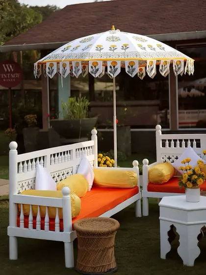 A vibrant Haldi seating area with traditional white wooden benches, colorful orange and yellow cushions, and custom-printed umbrellas.