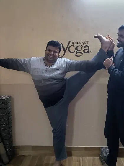 Progress, not perfection. A student smiles as he works on his standing split with wall support and instructor guidance, showing that advanced yoga is a journey accessible to all with the right approach.