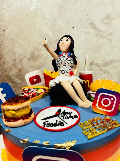 A close-up of a "foodie" and social media-themed cake, with a fondant figurine taking a selfie surrounded by edible burgers, fries, and social media logos.