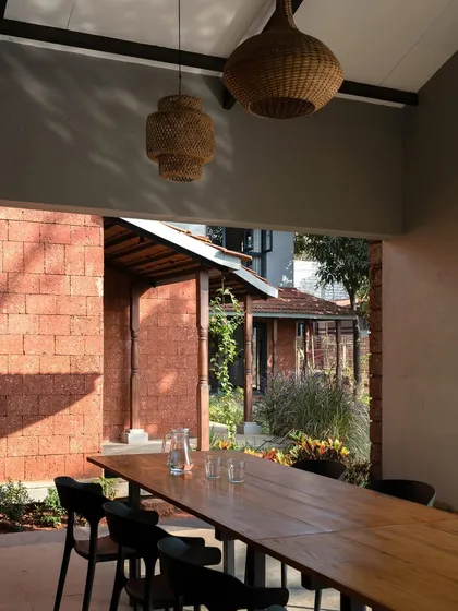The outdoor dining area at Maison Rouge is sheltered by a porch, looking out onto the laterite brick structures of the home. This space embodies the project's goal of creating a vacation oasis using local, vernacular building materials.