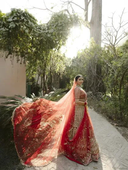 A stunning bridal portrait where the bride's long, red veil catches the sunlight. The lush green background and the golden light create a warm and ethereal image.