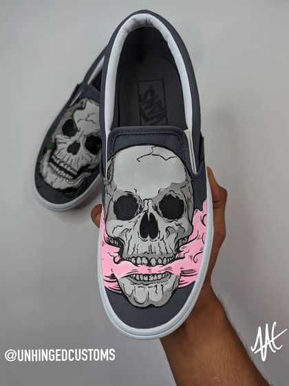 A dark and edgy skull design on a pair of Vans Slip-Ons. This side features a skull with pink smoke coming from its mouth.