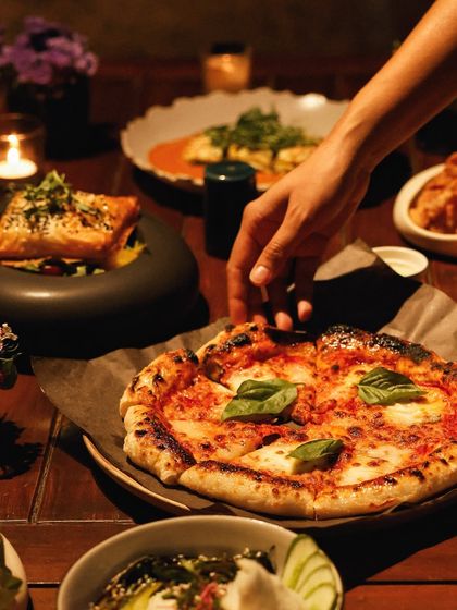 An intimate dinner scene, where a hand reaches for a slice of our freshly baked artisanal pizza, topped with basil and mozzarella.