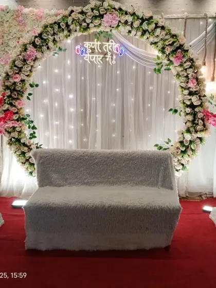 A beautiful floral setup for a traditional Marathi baby shower. The backdrop features a white curtain with fairy lights, a floral ring, and a sign that reads 'Kunitari Yenar Ga!', which means 'Someone is coming!'.