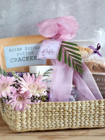 A beautiful woven basket for Women's Day, filled with healthy treats like Asian Spiced Puffed Crackers, wholewheat bread, and a fresh flower bouquet.