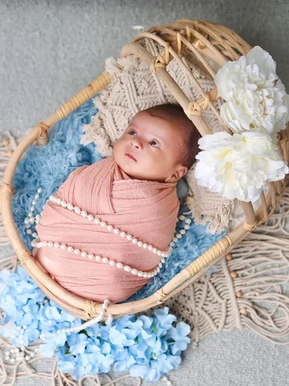 An awake and expressive newborn in a peach swaddle, looking up from a cane bassinet. The combination of blue and peach tones is soft and beautiful.
