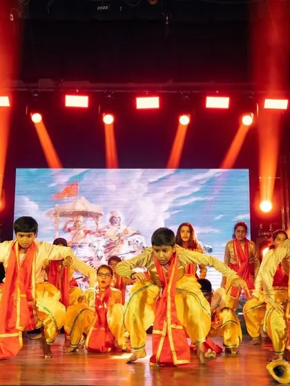 A group of young dancers in traditional attire on stage. The intense red spotlights from above highlight their performance, creating a visually striking scene.