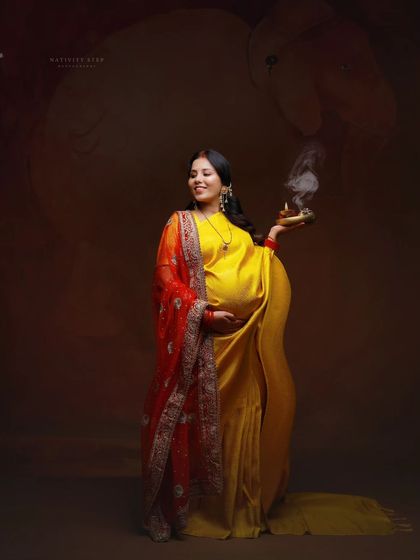 A vibrant and joyful portrait. The mother-to-be in a yellow saree with a red dupatta holds a diya, symbolizing auspicious beginnings.