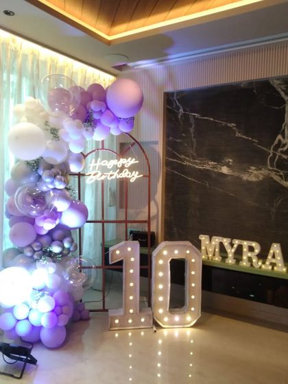 A sophisticated yet fun decoration for Myra's 10th birthday. The setup includes a window-pane style backdrop, a purple and white balloon arrangement, and large marquee numbers.