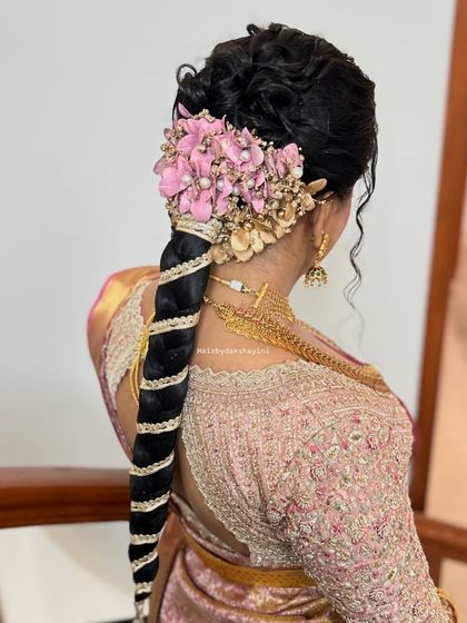 A very elaborate bridal hairstyle with a large floral arrangement of pink and gold flowers on top of a traditional braid wrapped in gold ribbon.